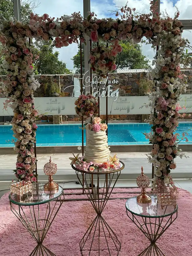Arco de flores rosas frente a la piscina y una torta blanca, armados para un cumpleaños